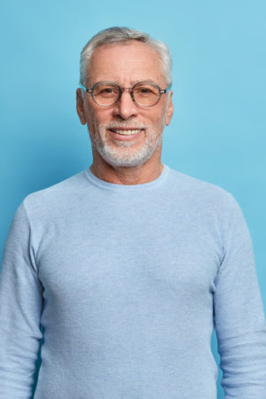 portrait-handsome-bearded-european-man-with-grey-hair-beard-smiles-pleasantly-looks-directly-front-being-good-mood-has-lucky-day-wears-spectacles-sweater-isolated-blue-wall