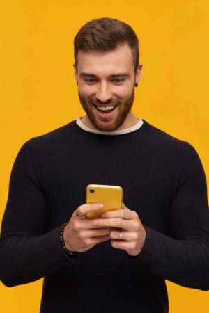Excited looking male, handsome guy with brunette hair and beard. Has piercing. Wearing black sweater. Holds smartphone and watching at it. Reading a message. Stand isolated over yellow background