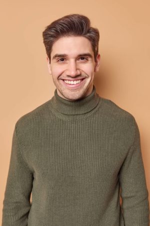 Portrait of handsome young man with dark hair smiles happily expresses positive emotions dressed in casual turtleneck hears good nes isolated over brown background glad to meet old best friend